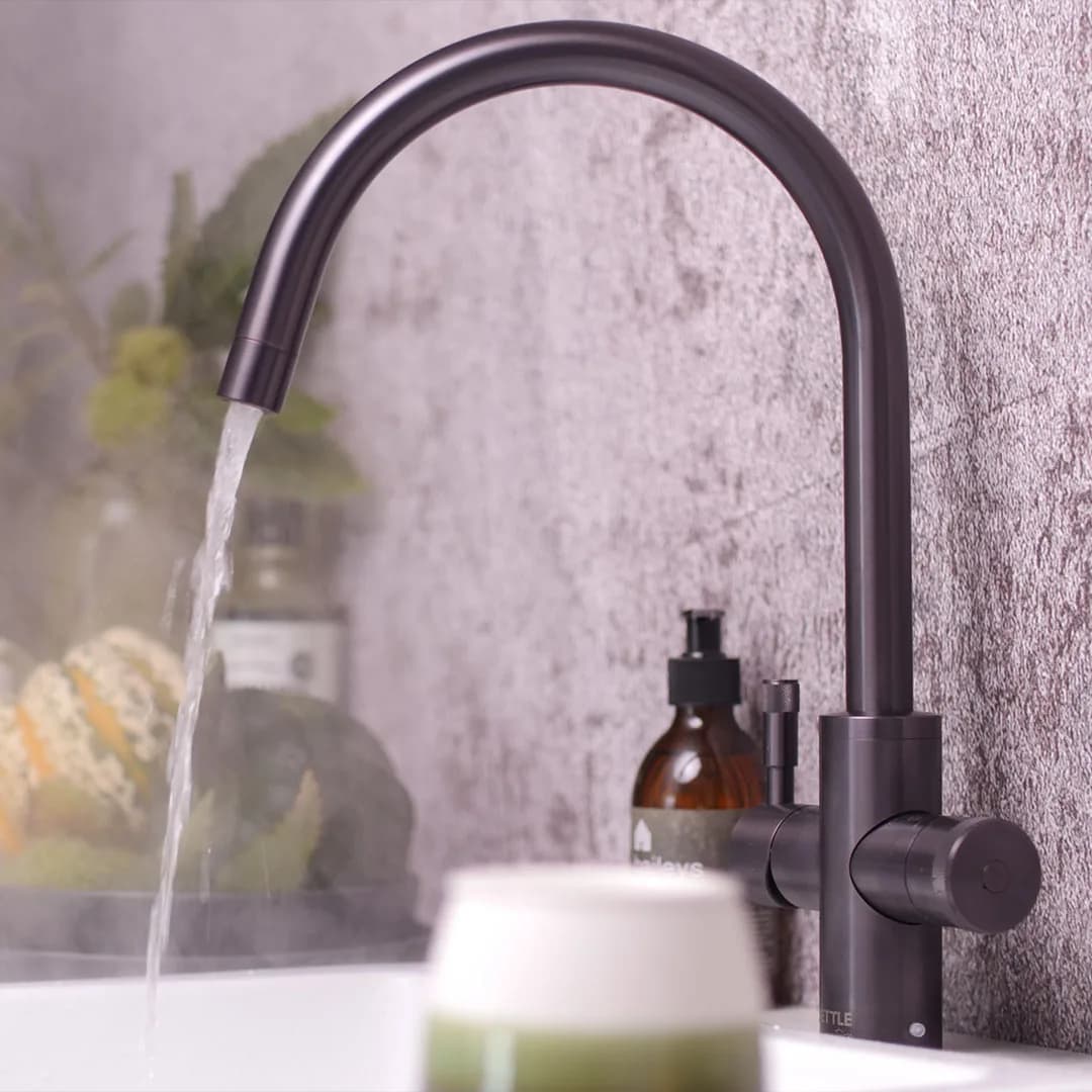 A gunmetal boiling water tap pouring into a sink beside a plant